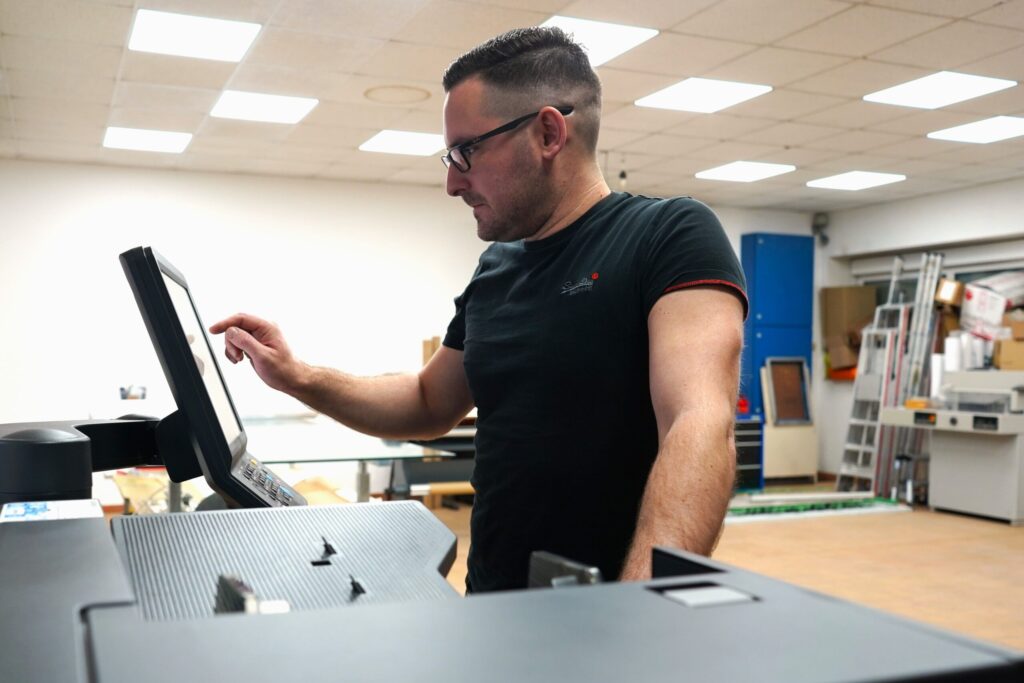 Mitarbeiter bedient einen großen Touchscreen-Drucker in einer Werkstatt. Im Hintergrund sind Regale, Kartons und Arbeitsmaterialien zu sehen.