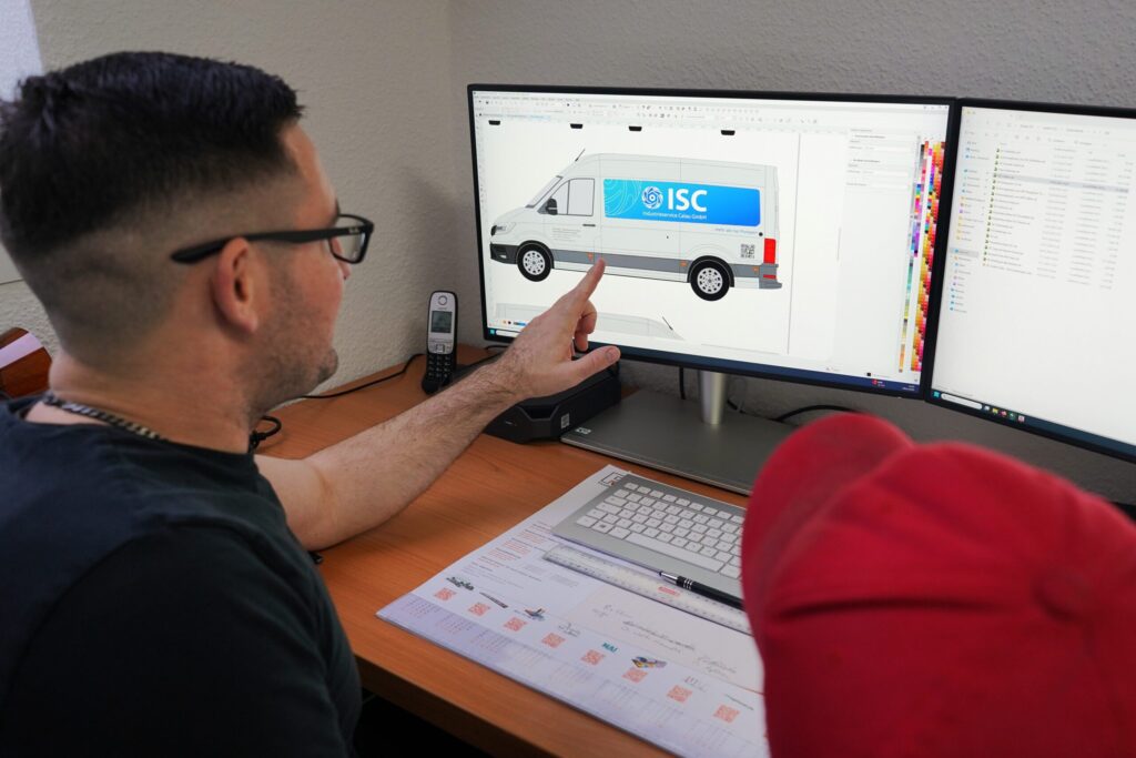 Mitarbeiter sieht sich am Schreibtisch eine Fahrzeugbeklebung am Computerbildschirm an. Er zeigt auf die Grafik eines Logos auf einem Transporter.