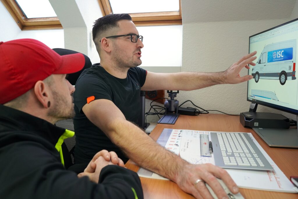 Zwei Männer besprechen am Schreibtisch eine Fahrzeugbeklebung am Computerbildschirm. Einer der Männer zeigt auf die Grafik eines Transporters.