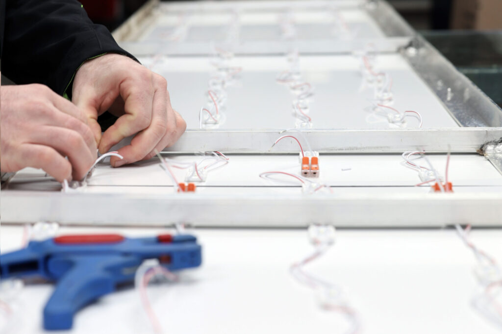 Nahaufnahme von Händen, die elektrische Kabel an einem LED-Modulrahmen anschließen, im Vordergrund liegt ein blaues Abisolierwerkzeug.
